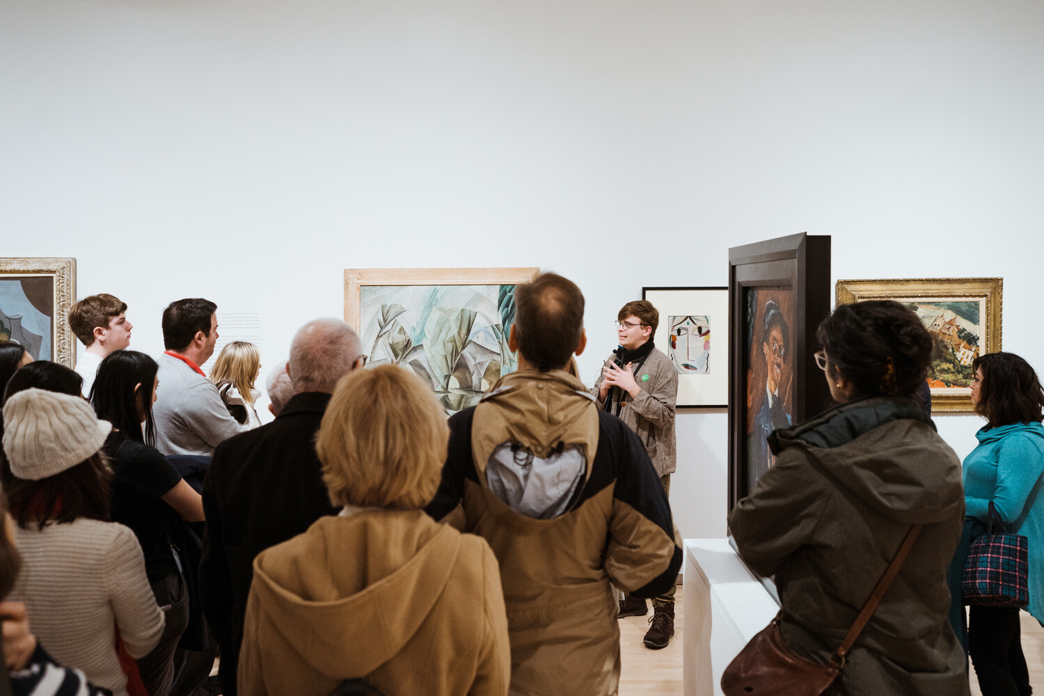 A person speaks to a large group of people in a gallery displaying paintings